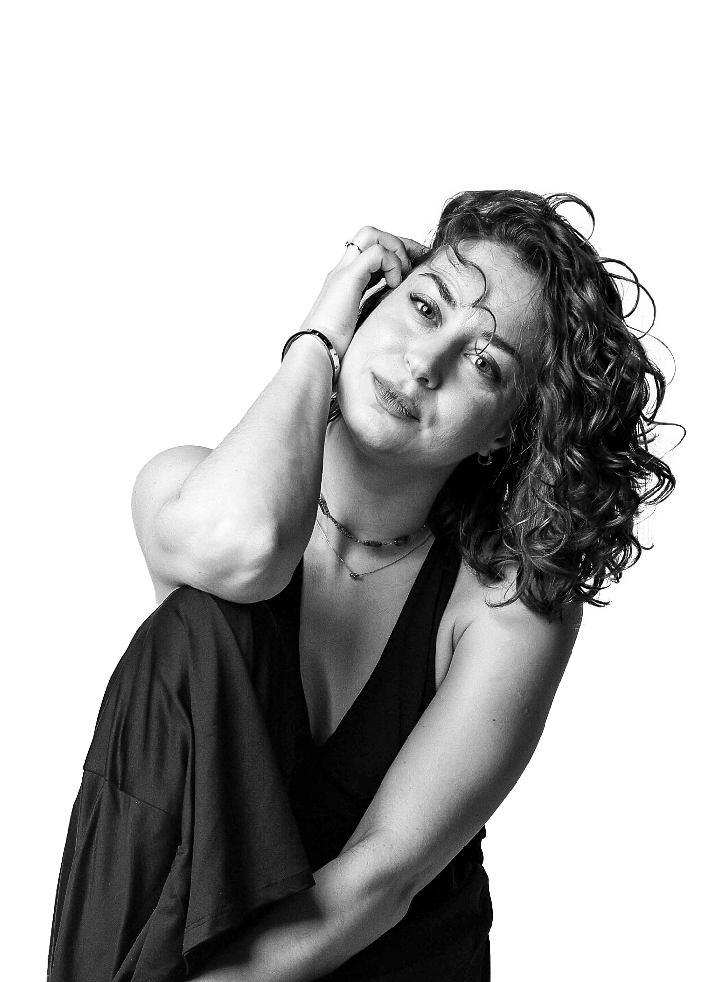 Schwarz-weißes, minimalistisches Portrait Shooting in Dresden mit junger Frau im schwarzen T-Shirt, die auf einem Hocker sitzt, lockiges Haar zur Seite trägt, eine Hand am Ohr hält und direkt in die Kamera blickt.